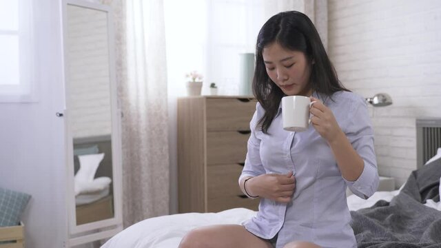 elegant Asian office lady touching her shirt and having a morning coffee drink in bed before she going to work.