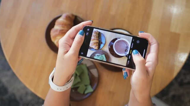 Close up of woman holds modern smartphone and taking pictures of her morning breakfast cocoa, green chocolate and tasty croissant to share photos on social media resources. Top view