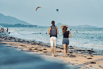 Young couple walks along the sandy beach at sunset in summer. Kite-surfings are riding in the background.