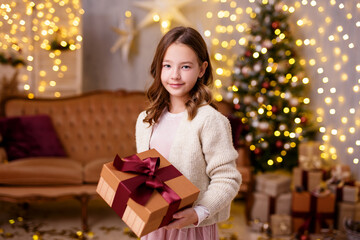 Fototapeta na wymiar Christmas and childhood concept - cute little girl holding gift box in decorated living room with Christmas tree and lights