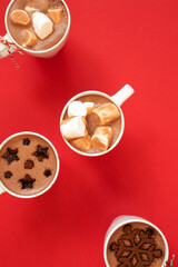 White cups with hot cocoa and marshmallows and spice cinnamon decor on red background, vertical. Winter holidays drinks concept