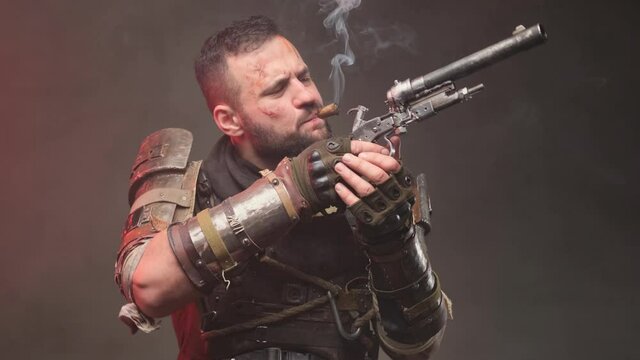 Bearded and dangerous military man in ragged armour examines pistol he holds and smokes cigar then aims with pistol and looks away in dark background.