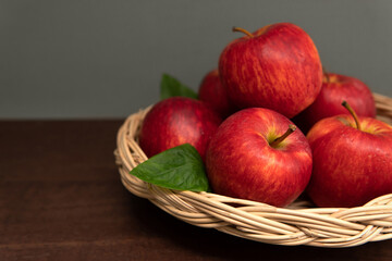 Closeup Fresh heap of red apple with leaves in a wicker basket on dark brown wooden table against grey color wall background