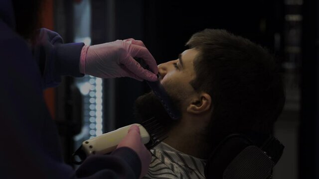 A bearded man trims his beard in a barber shop. Barber smoothes hair and cuts with a trimer