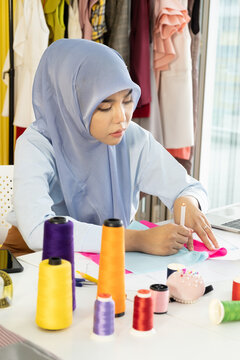 A young beautiful Asian Muslim woman designer who wore a hijab as a startup business owner is drawing the line on the colorful fabric on her working table.
