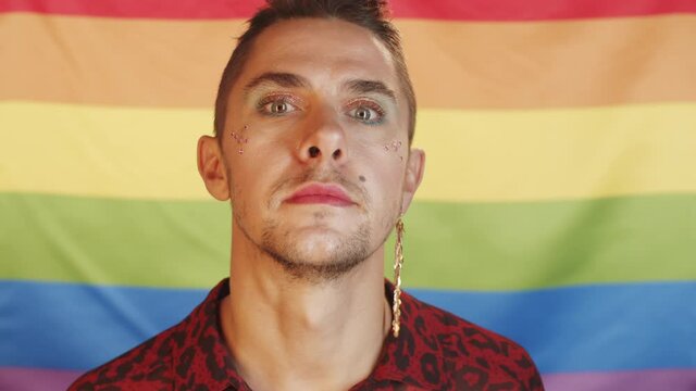 Portrait chest-up shot of young Caucasian gay man with makeup and trendy earring posing for camera against rainbow LGBT pride flag