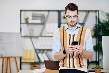 Pensive young businessman in glasses standing at his office desk and reading text messages on smartphone