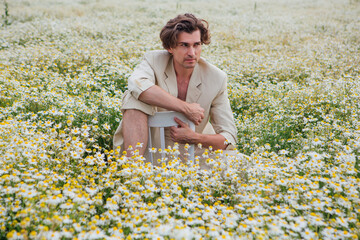 Tall handsome man sitting on a white chair in camomile flowers field