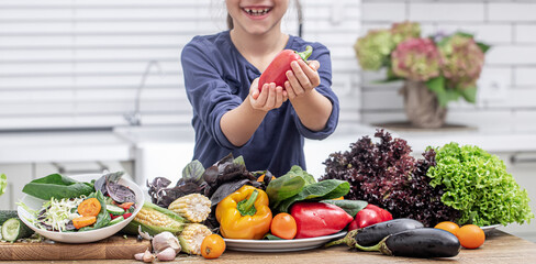 Little girl smiling and holding bell pepper on a background of many vegetables close up.