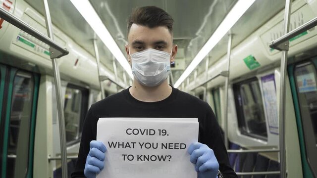 Young man in blue medical mask stand in underground carriage with "COVID 19. WHAT YOU NEED TO KNOW?" sign during pandemic covid-19 coronavirus quarantine. Keep yourself and others safety.