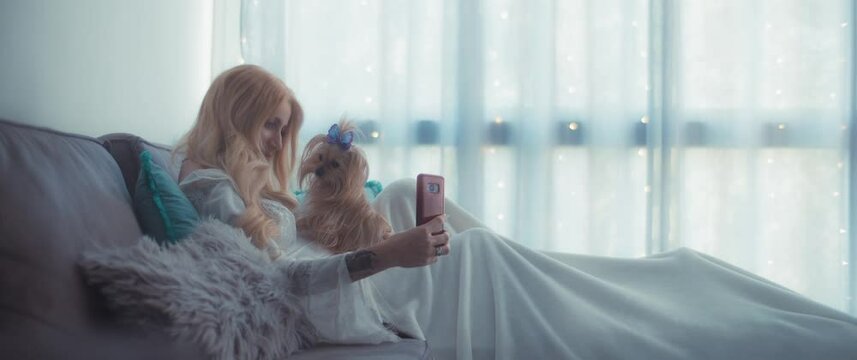 A pregnant woman taking a selfie with her small terrier puppy while sitting on a cozy couch. Slow motion, shallow depth of field, daylight, medium shot.