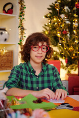 Portrait of smiling teenage boy in plastic glasses making Christmas decorations out of colored paper