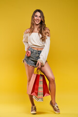 Beautiful young blonde woman smiling and holding shopping bags on a yellow background.