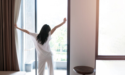 Girl opening curtains in a bedroom while stretching in the morning after getting up. Young woman standing in front of the bedroom window looking outside in morning sun.