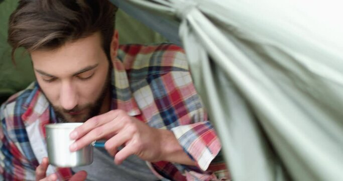 Close up of young man drinking hot coffee after hiking. Beautiful man relaxing in a tent. Camping passion