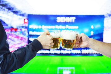 Two hands horizontal holding lager beer glass and clinking on background of football game. Sport fans cheer up. Friends leisure lifestyle concept.