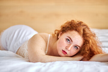 lovely young woman with natural red hair posing at camera, lying on bed alone, coquette female in...
