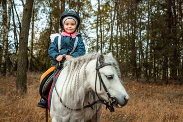 Fototapeta premium Little girl on a white pony on a background of nature. Jockey, hippodrome, horseback riding.