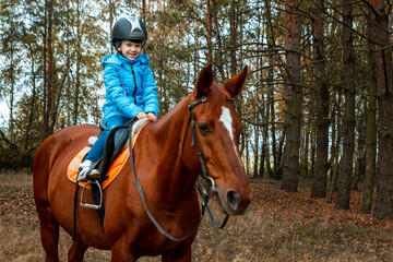 Fototapeta premium Little girl on an adult brown horse on the background of nature. Jockey, epodrome, horseback riding.
