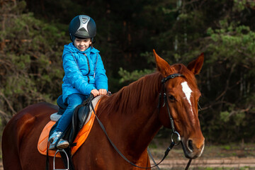 Fototapeta premium Little girl on an adult brown horse on the background of nature. Jockey, epodrome, horseback riding.