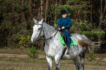 Little boy in a jockey cap on a white adult horse on a background of nature. Jockey, hippodrome, horseback riding