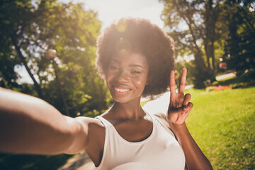 Photo portrait of shiny woman shooting selfie in sunshine making v-sign outdoors