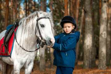 Fototapeta premium Boy in a jockey cap portrait, stands next to a white pony close-up on the background of nature. Jockey, epodrome, horseback riding.