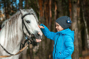 Obraz premium Little girl portrait, stands next to a white pony close-up on the background of nature. Jockey, epodrome, horseback riding