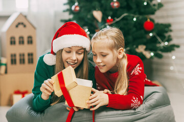 Two happy and beautiful teenage girls in New Year's outfits open gifts at home under the Christmas tree. Christmas mood. Concept for New Years holiday at home.