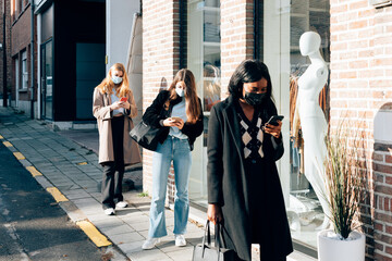 girls wearing face mask using a mobile phone while waiting on the street the opening of fashion...