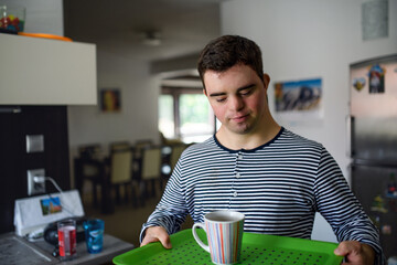 Down syndrome adult man standing indoors in kitchen at home, carrying cup on tray.