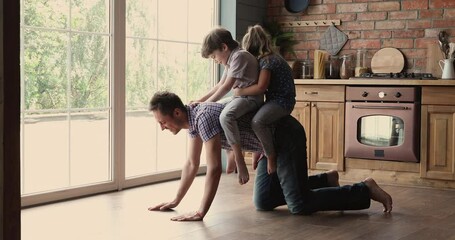 Little children sitting on daddy back riding having fun play together in kitchen. Dad crawling on warm wooden floor enjoy leisure funny time with pretty son and daughter, leisure, modern house concept