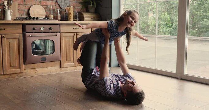 Dad lying on warm wooden floor in modern kitchen with underfloor heat system play with little daughter lifts up her on stretched arms kid imagines herself like plane fly in air, fun at home concept