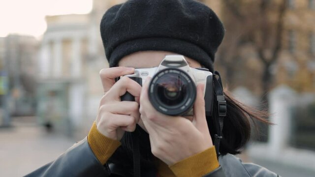 Young woman with photo camera looking straight, doing pictures close up. Beautiful asian woman in black beret holding a camera shooting in the street