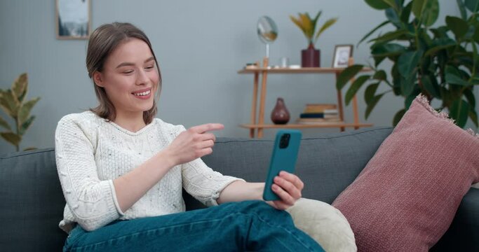 Happy woman showing with with deaf-mute sign language phrase I love you while sitting on couch. Pretty female person smiling and using smartphone while having video call. Hearing loss.