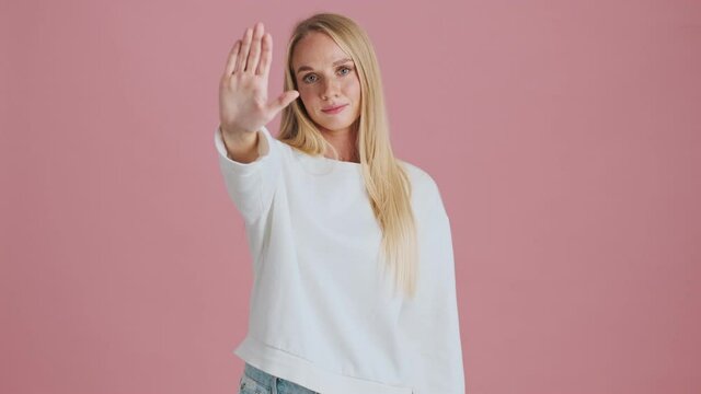 A serious young blonde woman is doing stop gesture standing isolated over pink background in studio