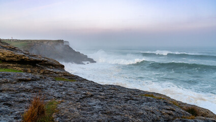 Fototapeta na wymiar storm waves crash onto the Cape Ajo in Spain with the lighthouse on the cliffs above at sunrise