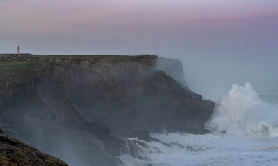 storm waves crash onto the Cape Ajo in Spain with the lighthouse on the cliffs above at sunrise