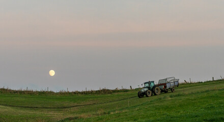 farmer with tractor and trailer cultivating fields under a full moon at sunset