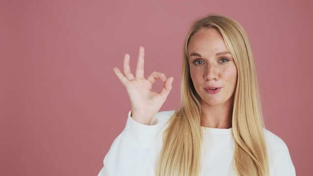A beautiful blonde woman is showing ok gesture standing isolated over pink background in studio