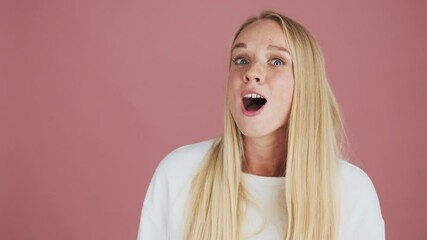 An attractive blonde woman is doing winner gesture standing isolated over pink background in studio