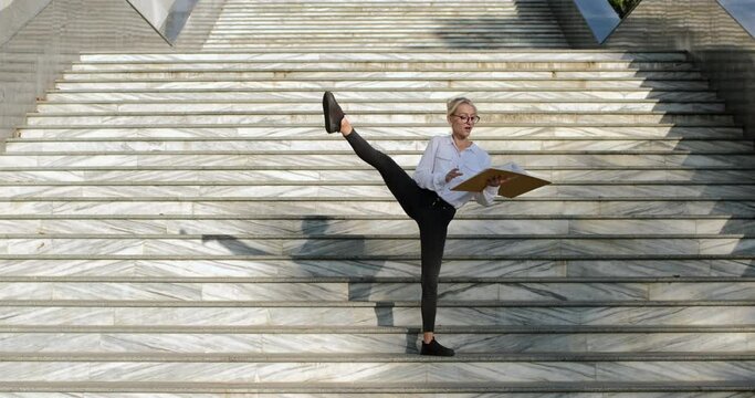 A beautiful girl, standing on the street stairs, revises documents in a yellow folder, lifts her right leg up. Flexibility in a work with papers and documents.