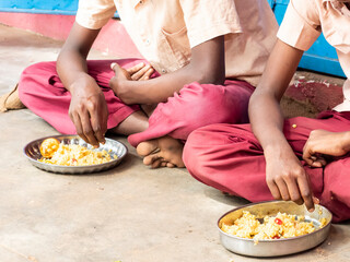 Unidentified poor classmates children with uniforms sitting on the floor outdoors, eating with...