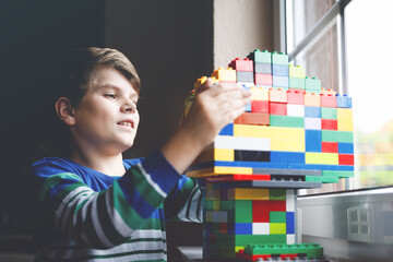 Little kid boy playing with lots of colorful plastic blocks. Adorable school child having fun with building and creating building by window. Creative leisure technic and robotic during corona time