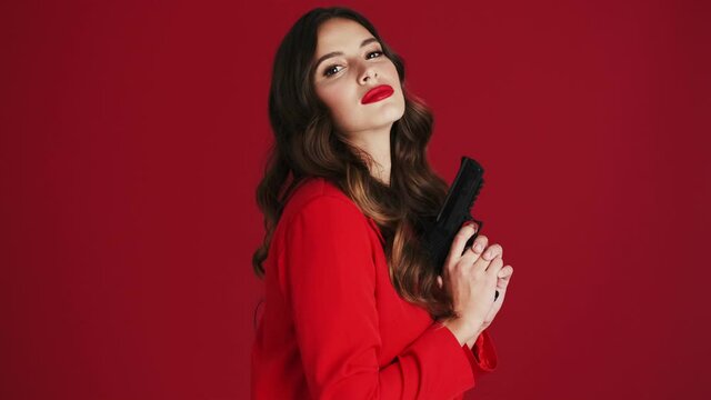 A beautiful woman in a red blazer is holding a gun standing isolated over red background in the studio