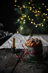 Christmas still life with homemade Panettone decorated as gift, blown candle and lights on pine branches on wooden table.