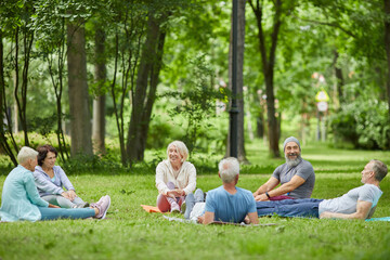 Wide shot of modern senior people spending summer morning together relaxing on grass in park after exercising