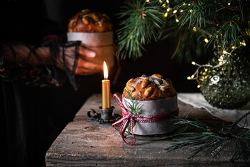 Christmas still life with homemade Panettone decorated as gift, vintage candleholder with lit candle and lights on pine branches on wooden table and woman holding panettone on background.