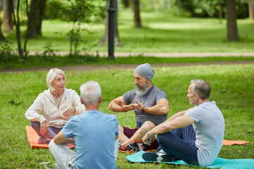 Fototapeta premium Group of active senior people gathered together in city park sitting on mats listening to their yoga trainer