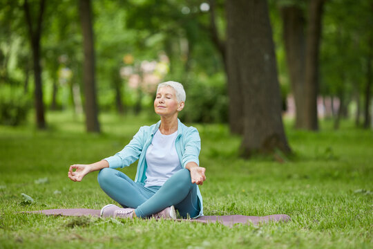 Beautiful senior woman spending summer morning time sitting on mat in park meditating with eyes closed